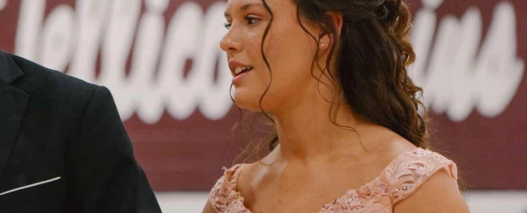 A woman in a pink dress with curly hair speaking at an event.