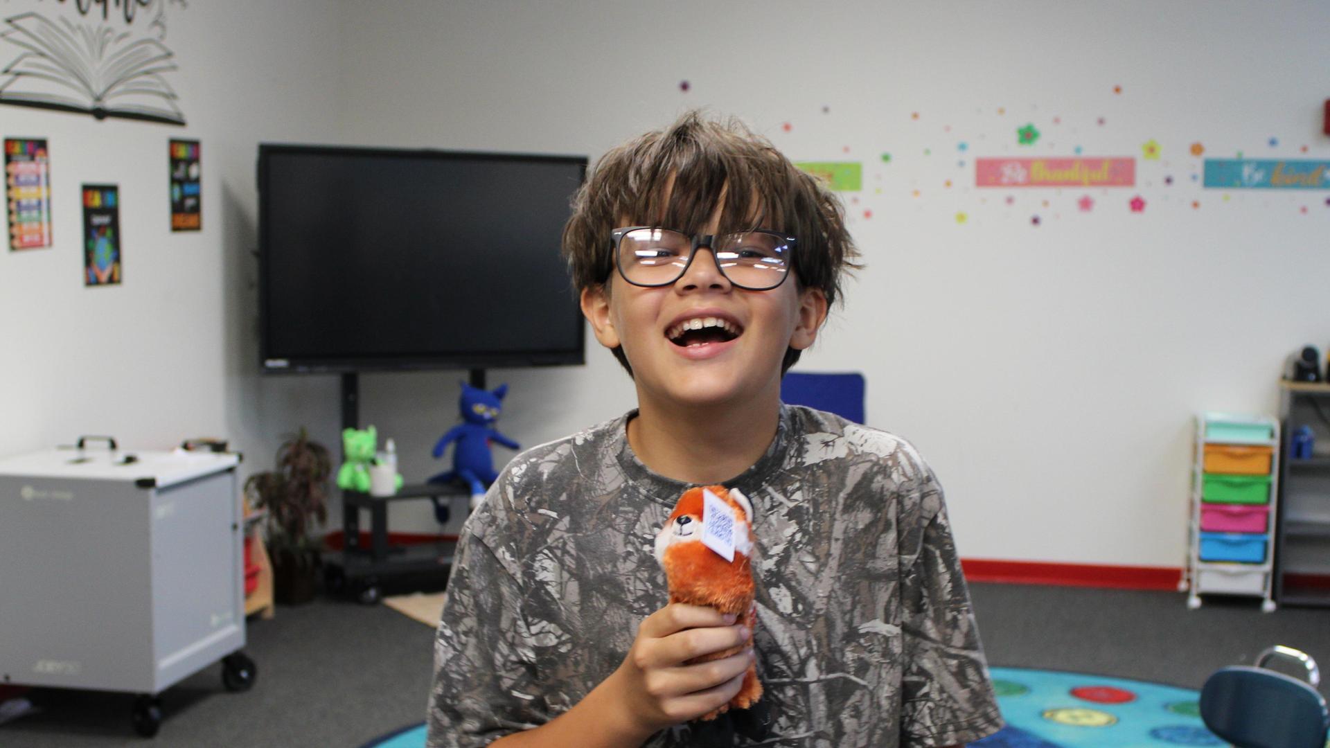 A boy laughing while holding a plush toy in a classroom environment.