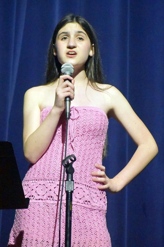 Girl in a pink dress singing into a microphone on stage.