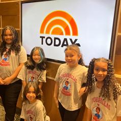 A group of five girls wearing matching shirts posing in front of a TV screen with 'TODAY'.