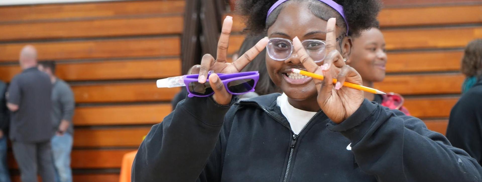 A smiling person making a peace sign, wearing glasses in a gym.