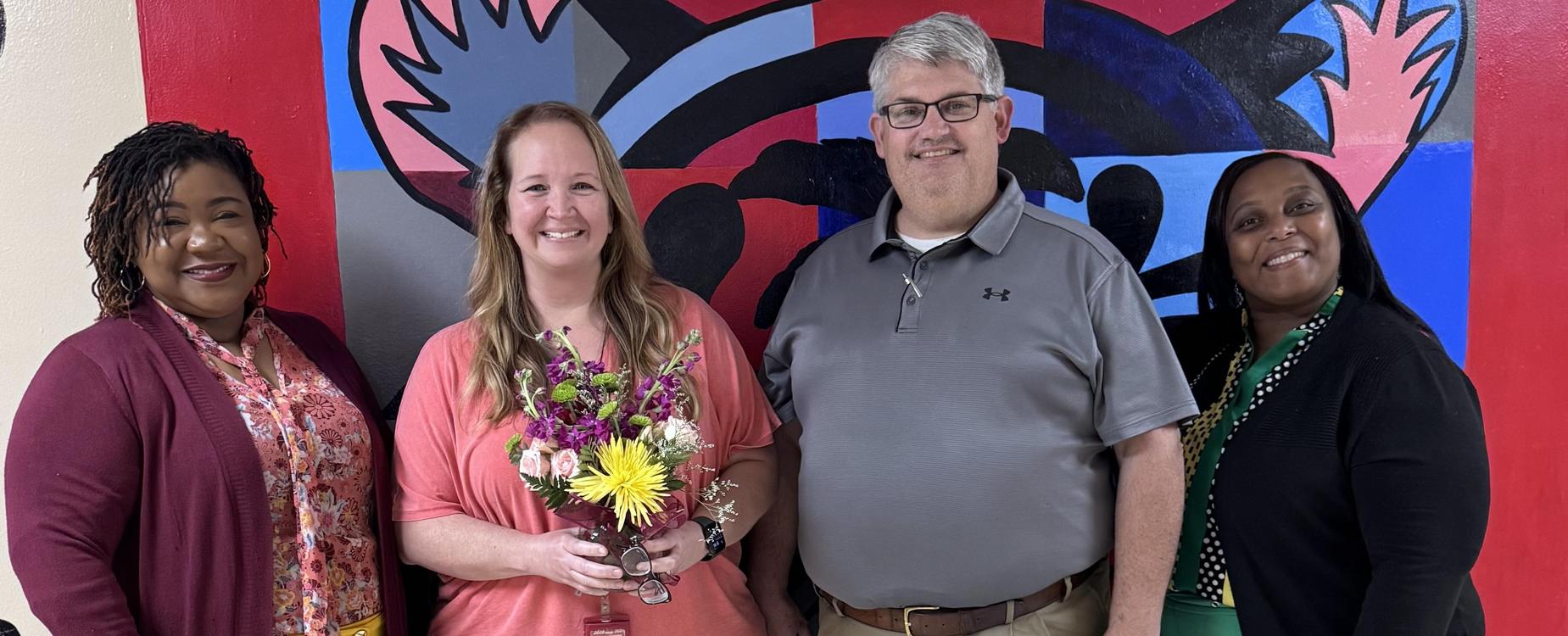 Four people holding flowers in front of a colorful mural backdrop.