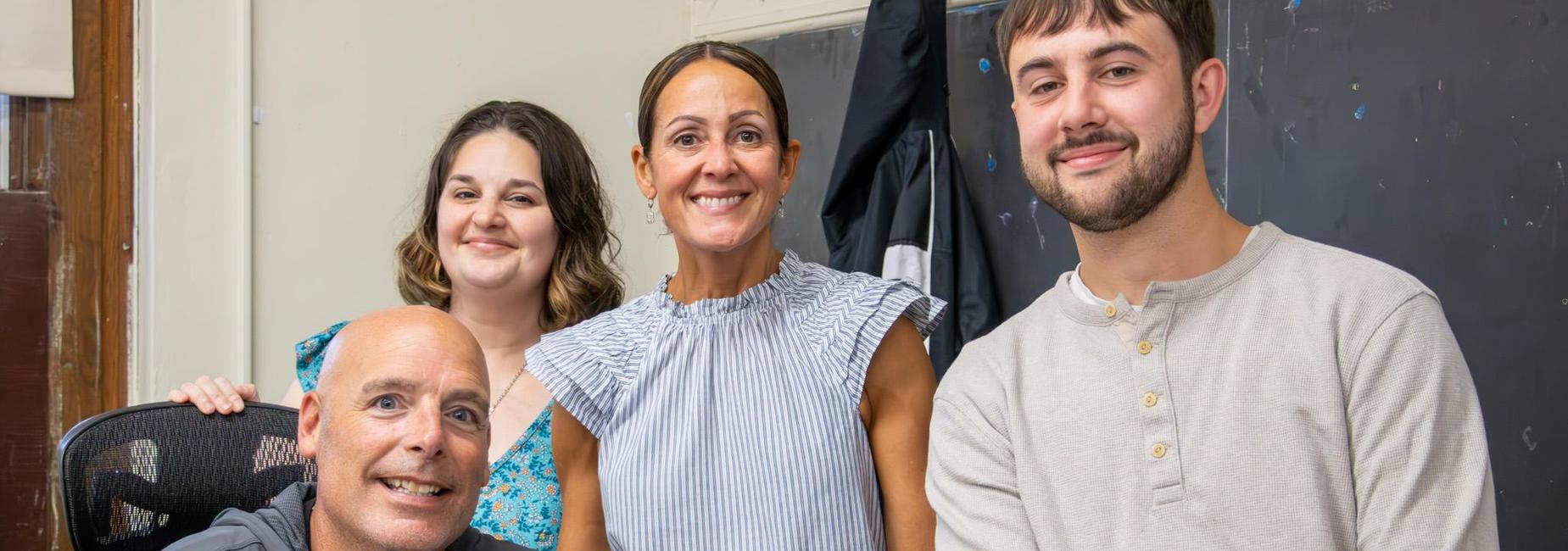 Four smiling individuals posing together in a classroom setting.