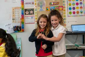 Two students at Arco Iris Spanish Immersion School hold their hands together to make a heart. They are in a colorful classroom.