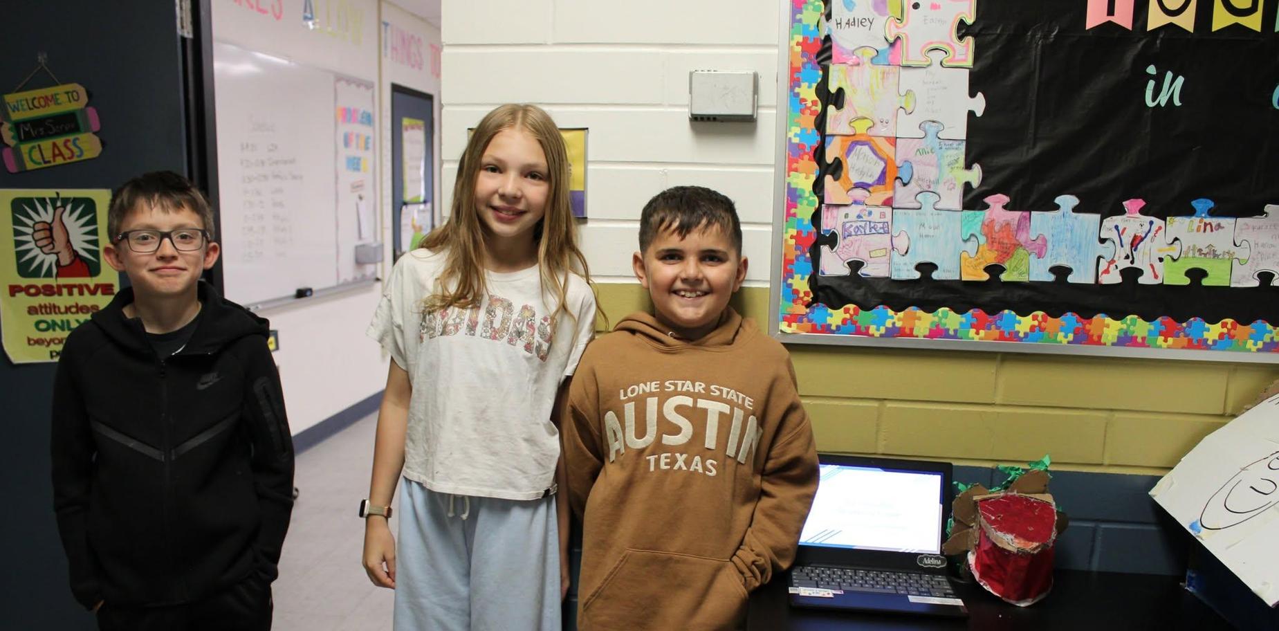 Three children pose together in a classroom with a laptop and project displayed.