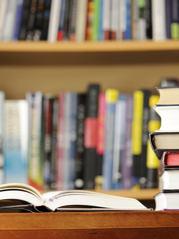 An open book in the foreground with stacked books and shelves in the background.