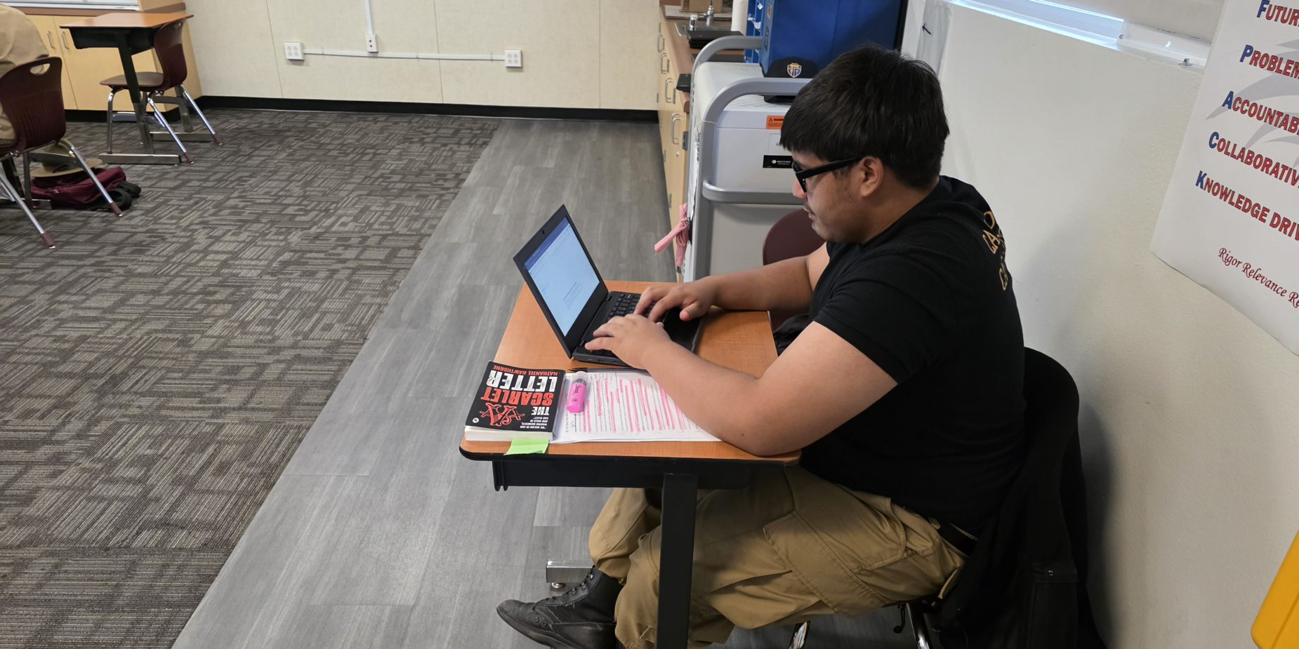 Student working on a laptop at a desk, with a notebook and textbook beside him.