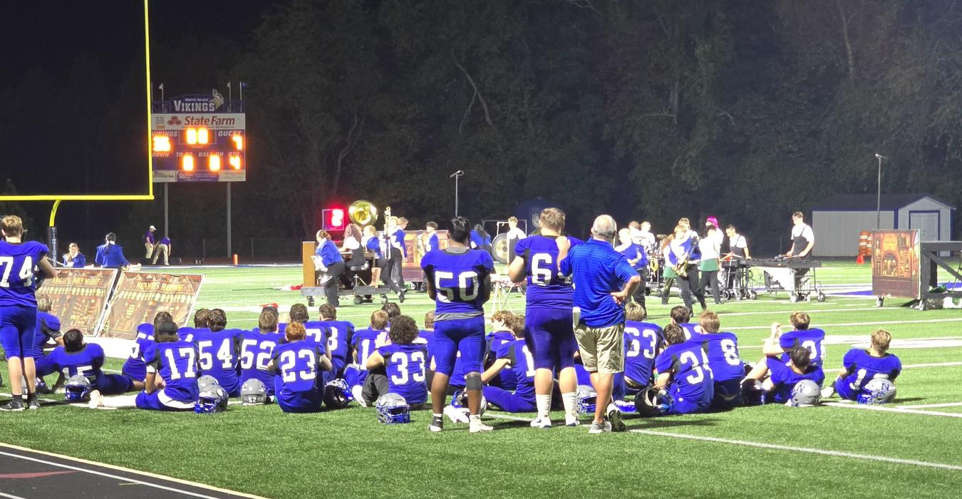 A group of football players sitting on a field, with a scoreboard in the background. Watching Vikings Marching Band perform.