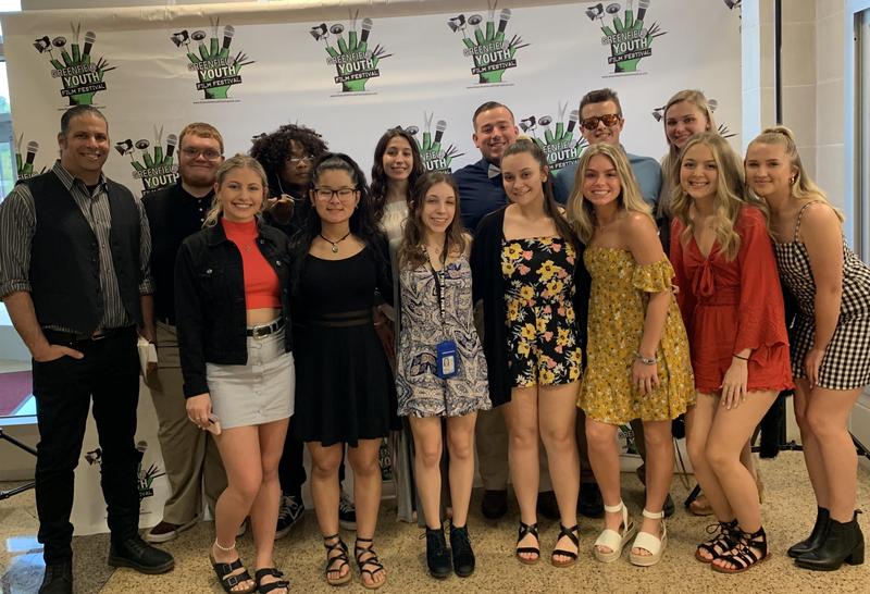 13 boys and girls along with their teacher standing in front of the Greenfield Youth Film Festival backdrop. These students competed in the film fest.