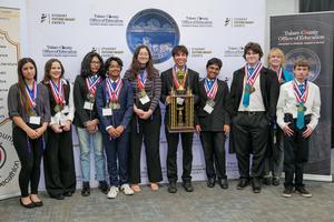 The Harmony Magnet Academy Academic Decathlon team stands proudly with their trophy and medals after winning the 2025 Tulare County Academic Decathlon. The students are smiling in front of a Tulare County Office of Education backdrop, showcasing their achievement with multiple medals around their necks and a large championship trophy held at the center.