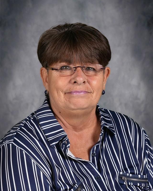 Smiling woman with short brown hair, wearing glasses and a yellow top, posing against a gray background.