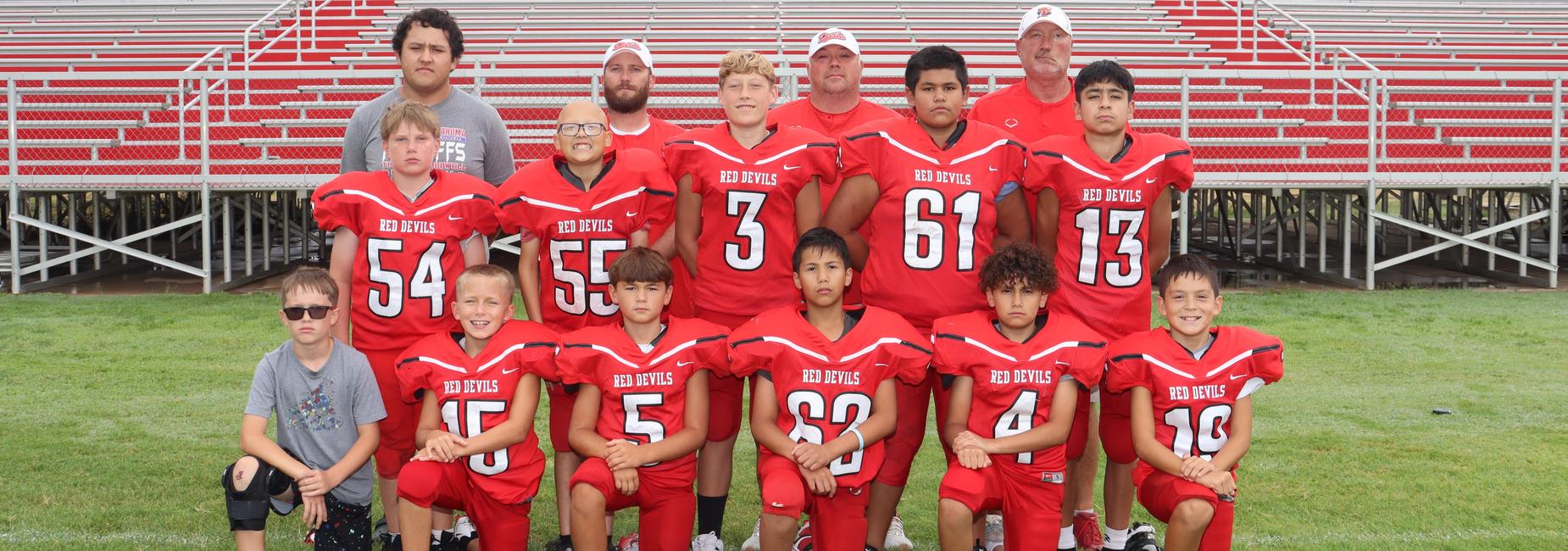 Youth football team posing together on a field, wearing red uniforms with a bleacher backdrop.