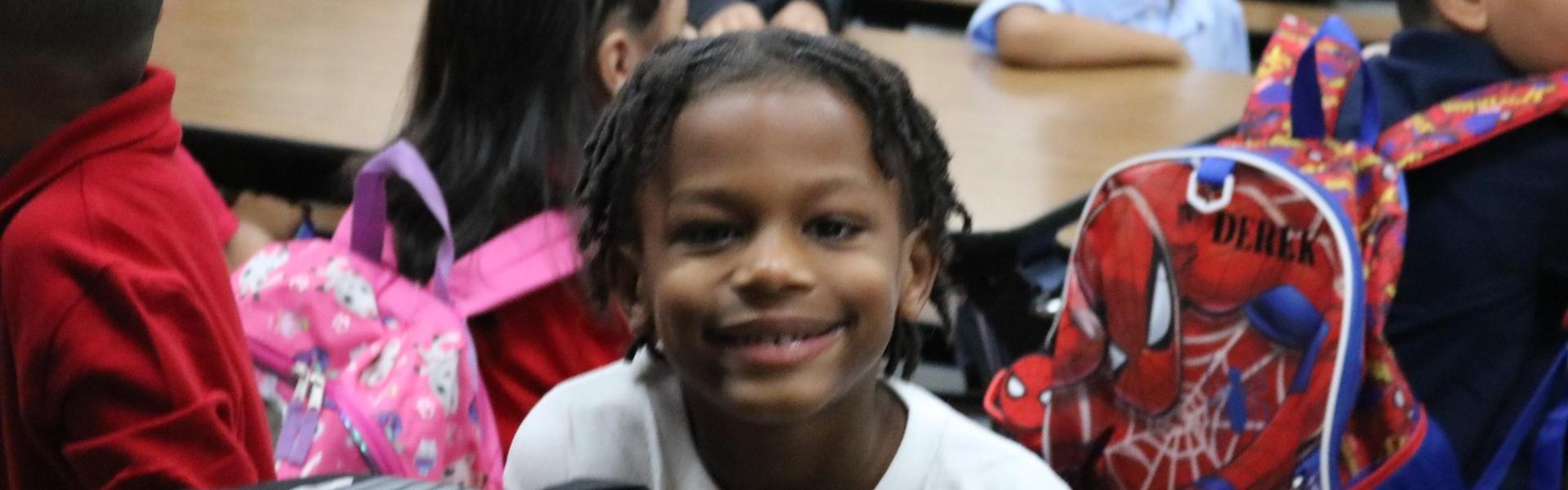 Smiling child with curly hair, sitting in a classroom with other students.