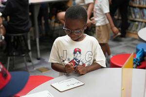 boy works on a paper while seated at a small white table