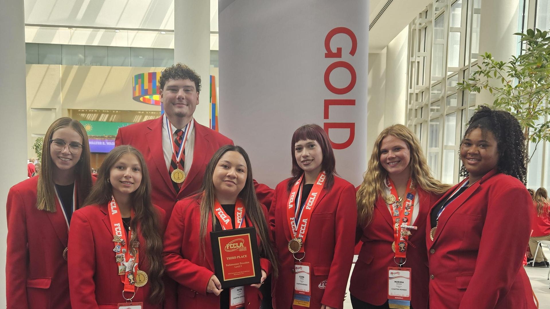 Group of seven students in red jackets holding an award and wearing medals.