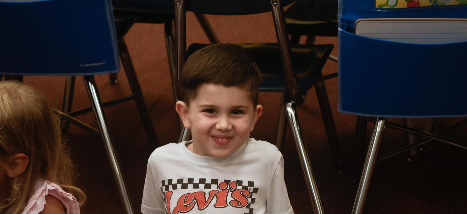 A smiling child in a T-shirt with a pizza graphic sits on a classroom floor.