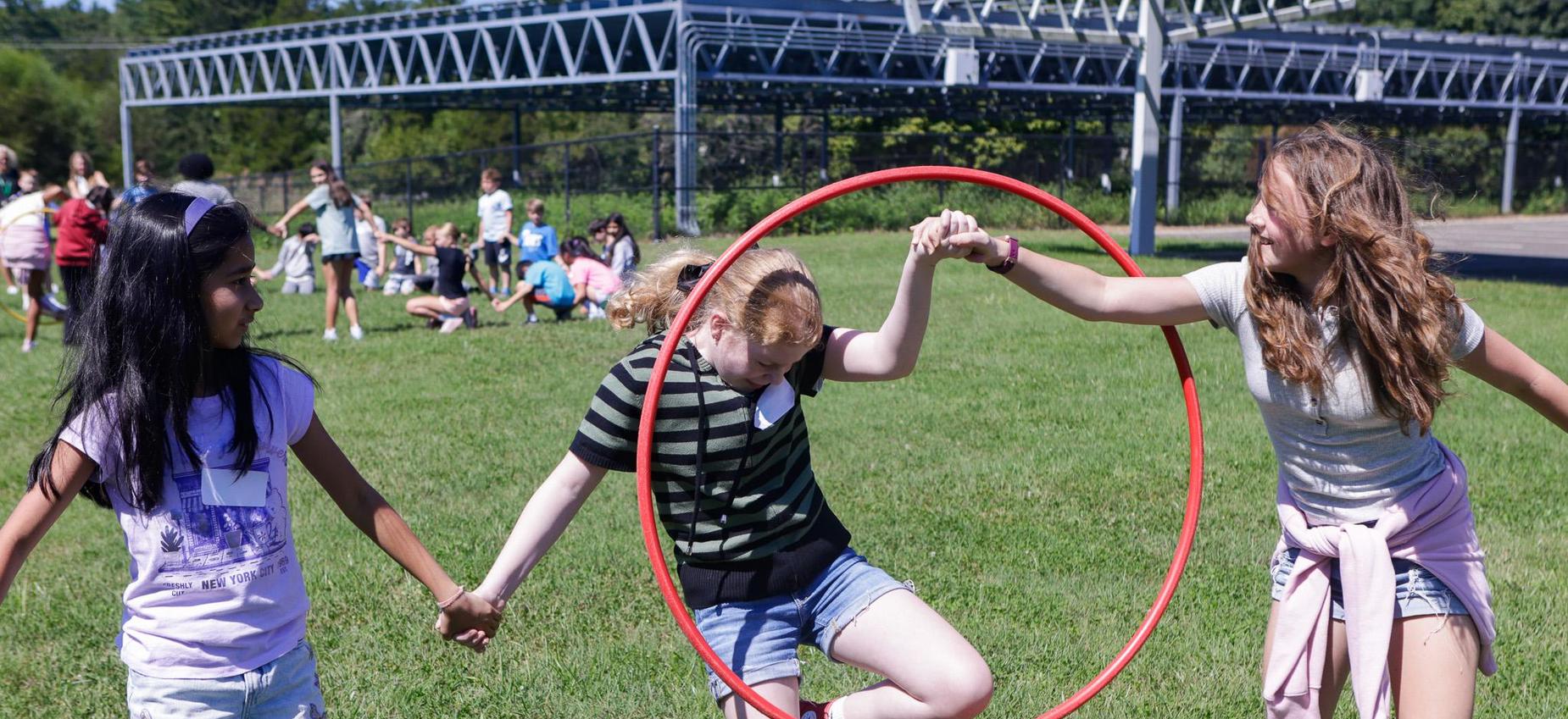 Three children playing together on grass, one stepping through a hoop.