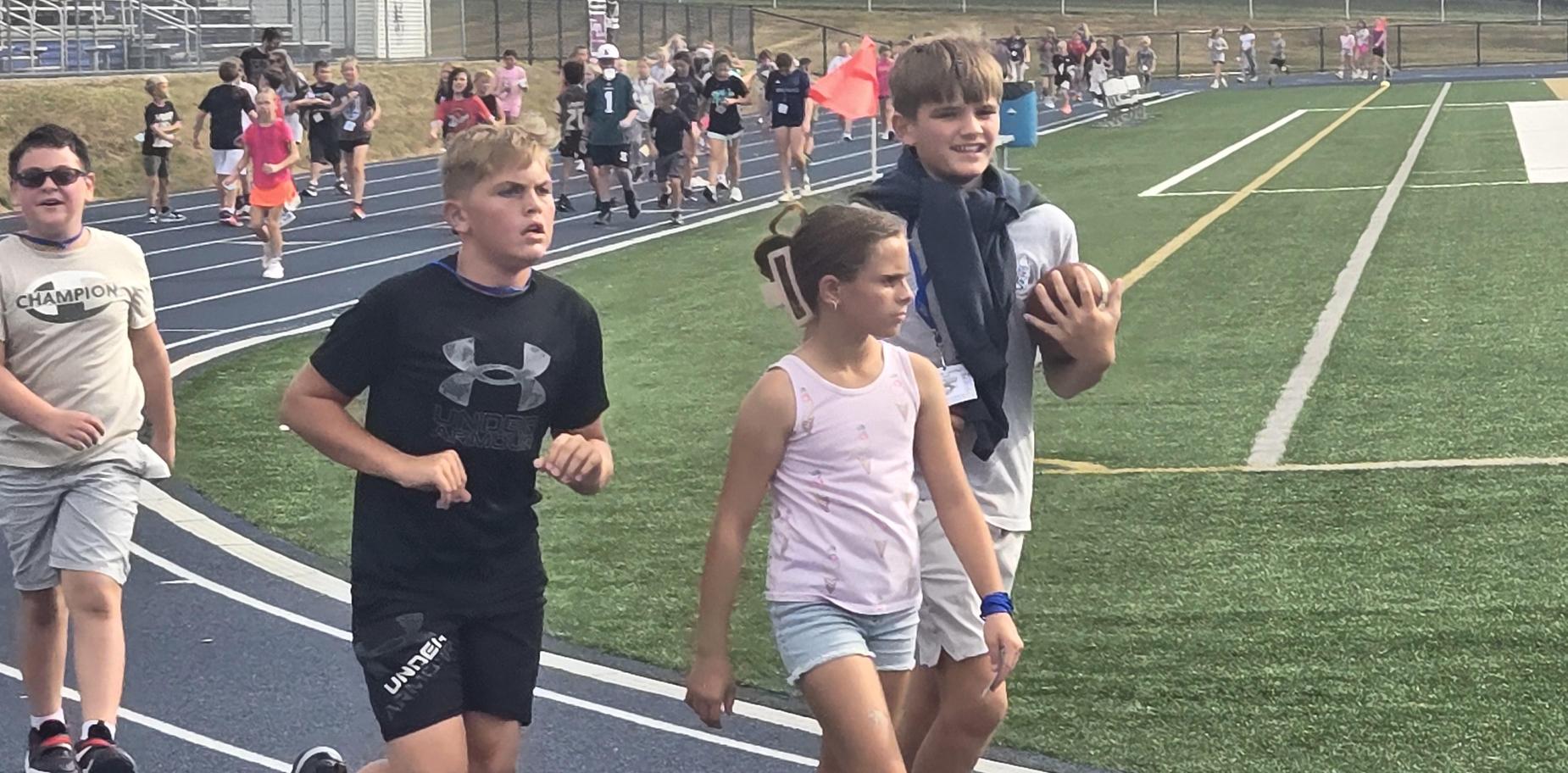 Boys and a girl walking on track, holding a football, with kids running behind them.