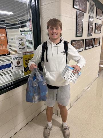 Smiling young man in a white hoodie holding a bag and a box in a school hallway.