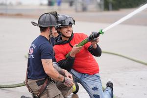 A smiling student in a red shirt and helmet operates a fire hose under the guidance of a firefighter wearing Porterville Fire Department gear. Water sprays from the hose as part of a hands-on fire training experience.