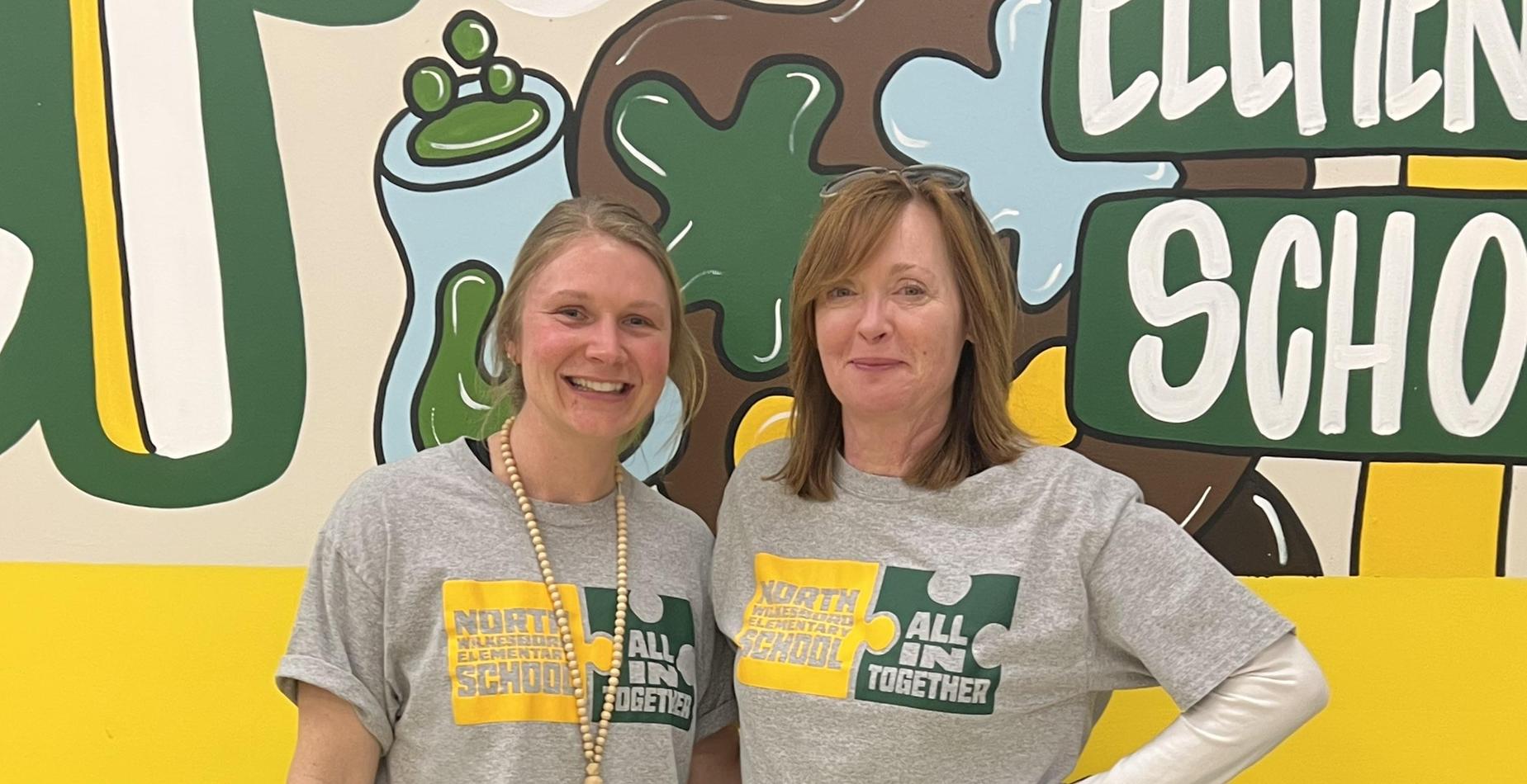 Two women in matching gray t-shirts with school logos, smiling together indoors.