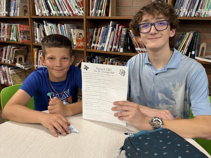 Two boys smile at the camera, one holding a paper, in a library setting.