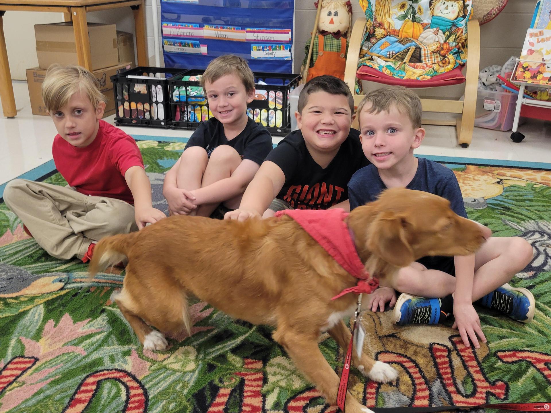 Four children laughing as a dog walks around them in a classroom on a patterned rug.