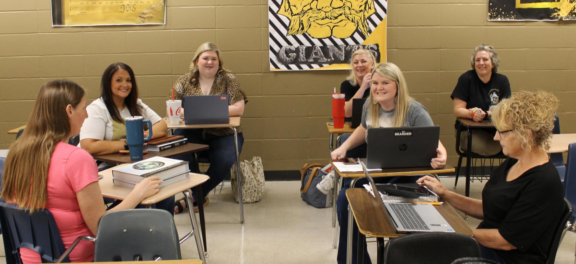 Seven individuals gathered in a classroom, engaging in discussions and working on laptops.
