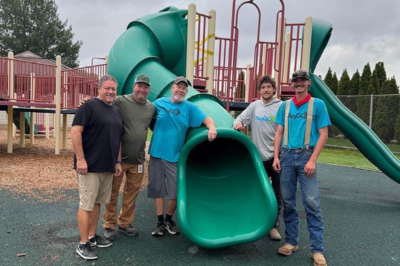 A green sliding board with five men standing alongside on a playground