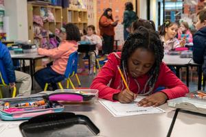A student at Arco Iris Spanish Immersion School works at a desk in a classroom. There are students and teachers in the background.