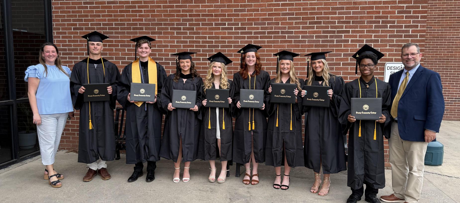 Graduates in caps and gowns holding diplomas, standing in front of a brick wall.