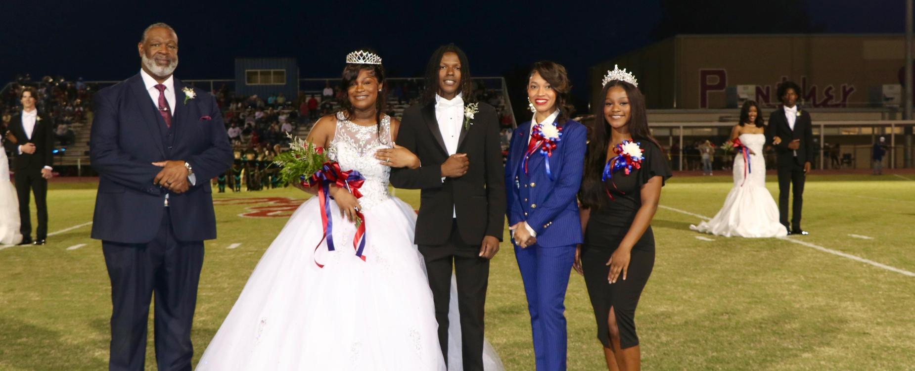 A group of five individuals, including a crowned queen and king, stand on a football field during an event.
