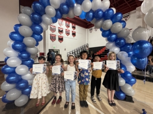 Smiling elementary students hold awards certificates under a balloon arch