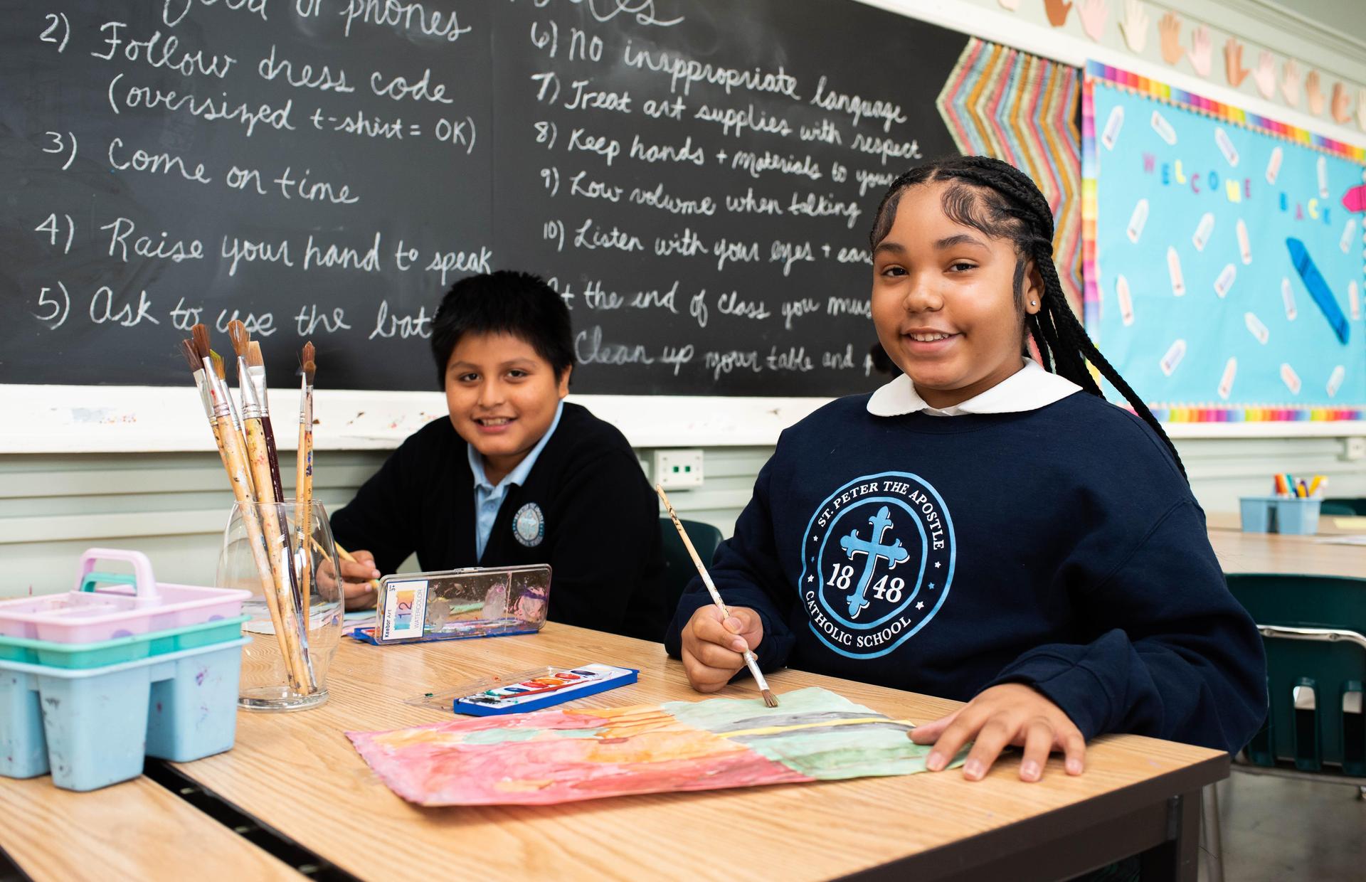 Two students create art at a classroom table, smiling and engaged in their work.