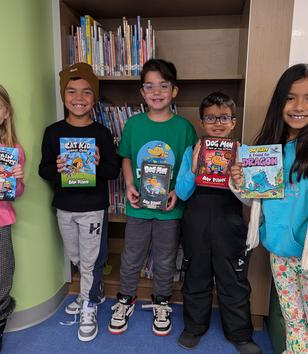 Five children in a library holding various graphic novels, smiling happily.