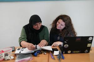 Two women collaborating on documents at a table, with stationery and a laptop.