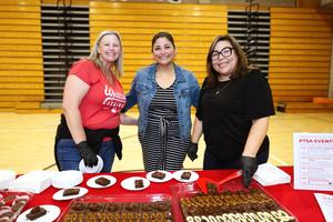 Three women smile for a photo while standing behind a table displaying brownies and other baked goods at a Whittier High School PTSA event in the school gymnasium.