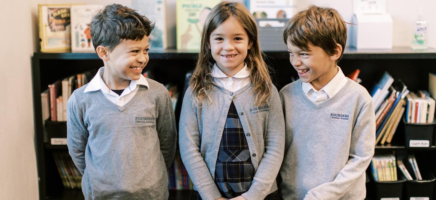Three smiling elementary students stand in front of a bookshelf