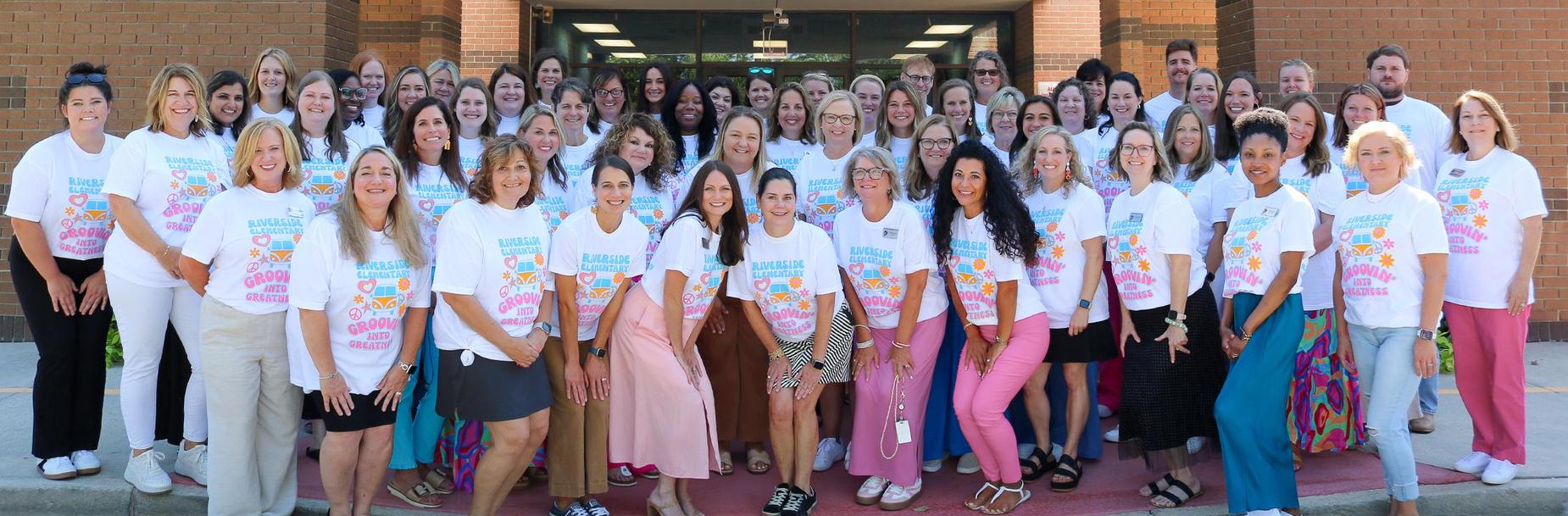 Group of school staff posing together in matching white shirts outside a building.