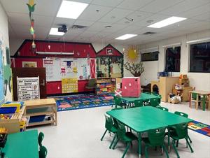 A bright classroom with a barn-shaped wall, small green tables, and a rug.