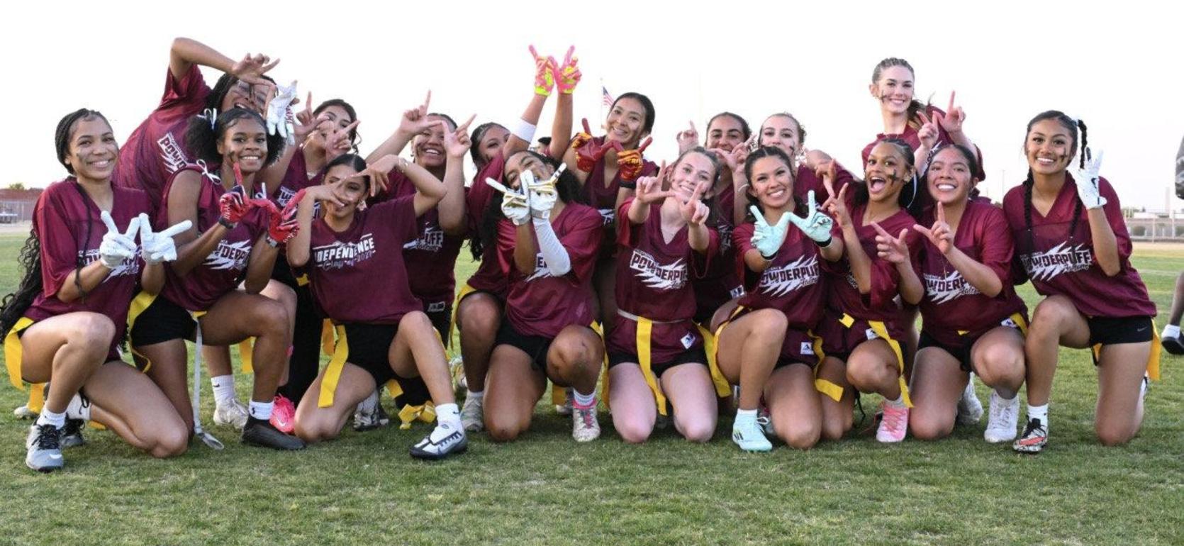 A large group of young women in sports gear celebrate with hand gestures on a field.