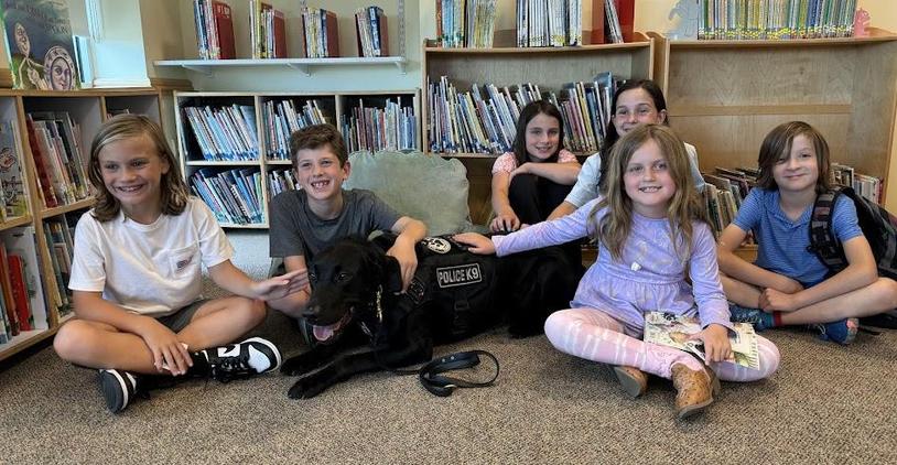 Six children sitting with a police dog in a library, smiling and interacting.