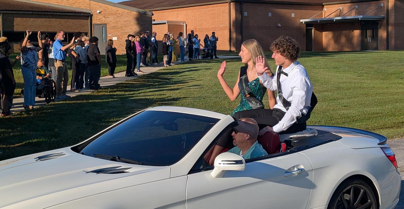 Two teenagers in formal attire wave from a convertible car as a crowd watches.
