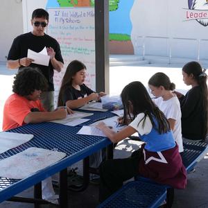 students coloring at a picnic table while a staff member reads to them