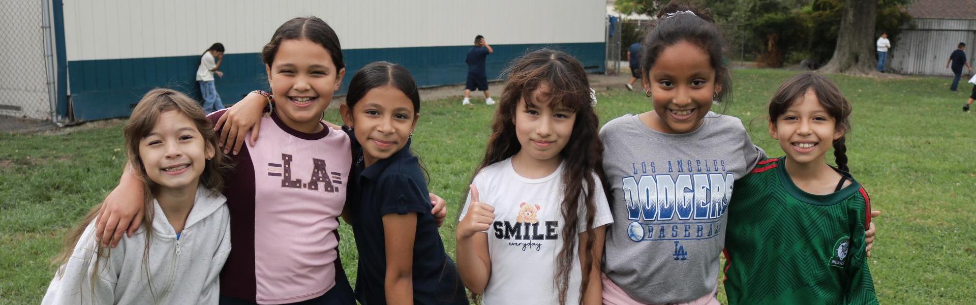 Group of six children smiling and posing outdoors on grass.