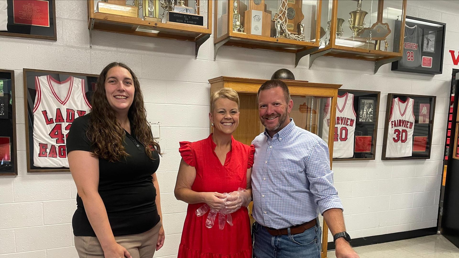 Three people smiling together in a school gym with sports jerseys in the background.