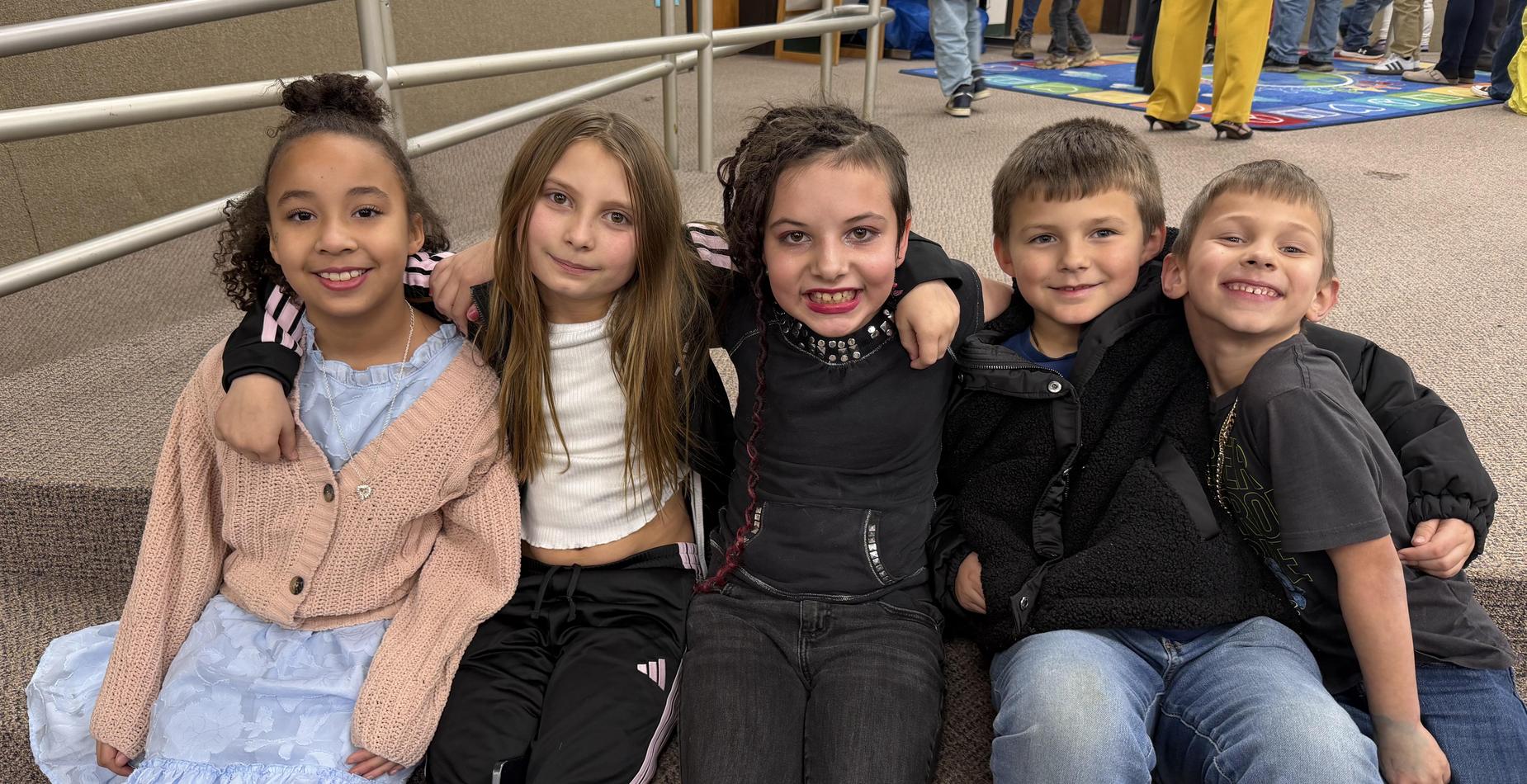 Five children sitting together, smiling and wearing casual clothing.