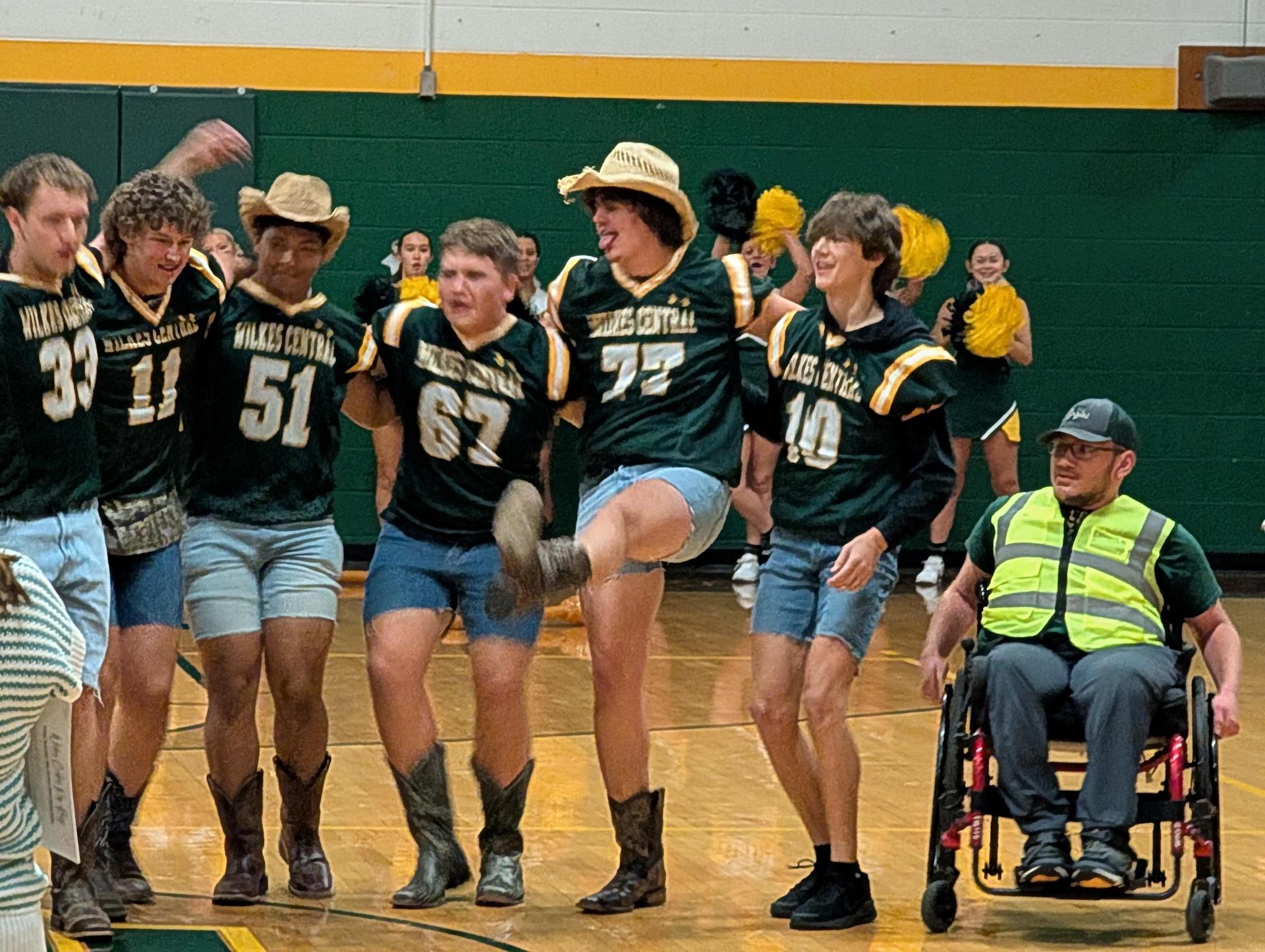 Group of male students in cowboy outfits performing a dance on a gym floor, with one in a wheelchair.