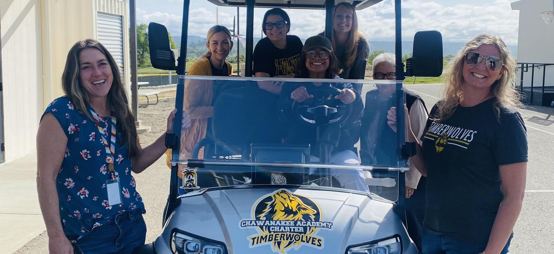 Group of smiling women and one man posing with a golf cart in daylight.