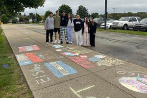 Group of six students posing beside colorful chalk drawings on a sidewalk.